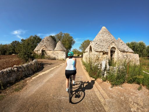 A back shot of an adult woman riding a bicycle on a lane through old trulli houses in Puglia, Italy