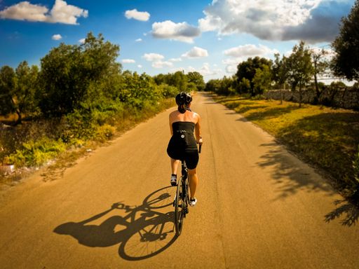 A back shot of an adult woman riding a bicycle on a lane through olive groves on a sunny day in Puglia, Italy