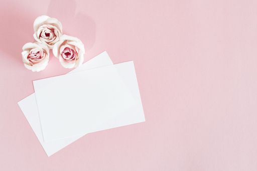 A top view of white invitation cards placed on a pink surface next to pink flowers