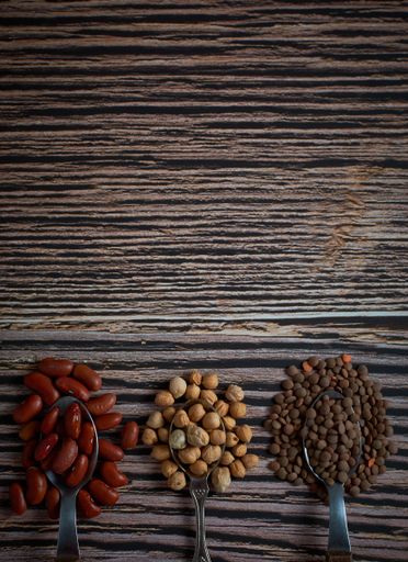 A top view vertical of three overflowing spoons of red kidney beans, chickpeas, and brown lentils on a wooden surface