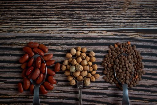 A top view closeup of three overflowing spoons of red kidney beans, chickpeas, and brown lentils on a wooden surface
