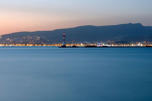 A horizontal long exposure shot of the sea with city at the background at night. A lighthouse in the center