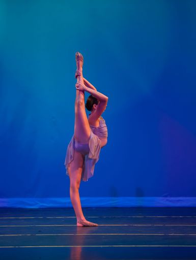 A vertical shot of a ballerina standing in a high kick pose on a stage with blue background