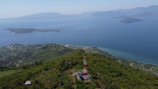 An aerial view of a telecommunication tower and solar panels on top of a forested mountain on the coast