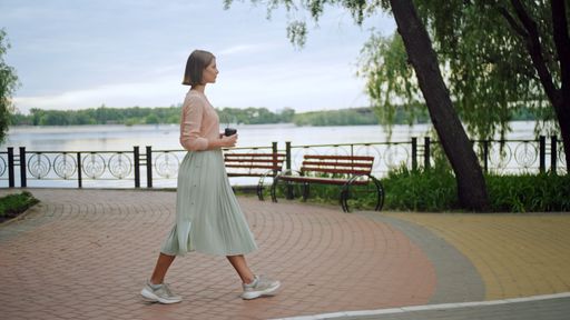 A young female in a pink shirt and long green skirt walking on a pier with a cup of coffee