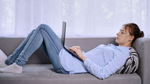 A young female in a striped shirt and jeans laying on the sofa using her laptop