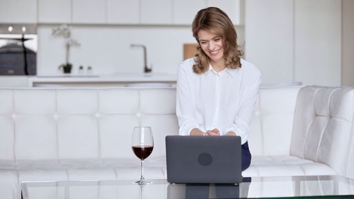 A young businesswoman in a white shirt sitting on a white sofa using her laptop near a glass of wine