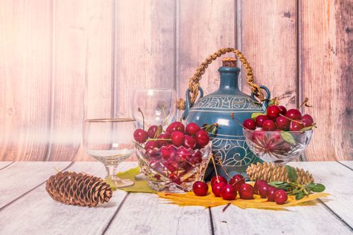On wooden table are vases with ripe cherries, earthenware jug of wine, wine glasses, yellow maple leaves and fir cones. Still life with festive mood.