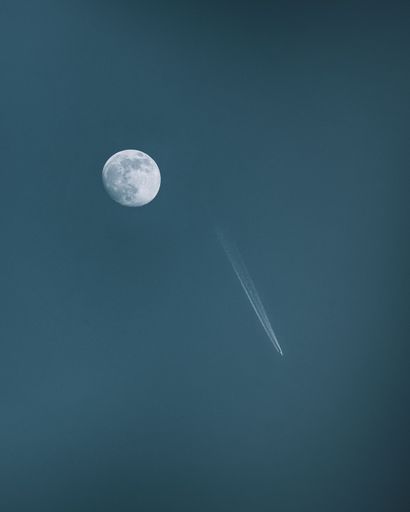 A vertical shot of a textured white full moon in a blue sky with a flying airplane