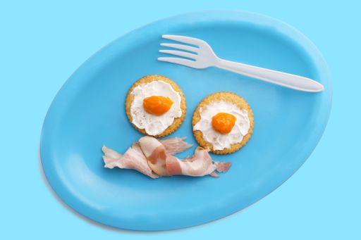 A top view of salty cracker with cheddar and cream cheese in form of egg next to bacon on blue plate and background