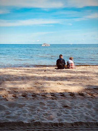 A vertical shot of a couple sitting on a sandy beach against blue Baltic sea in Gdynia, Poland
