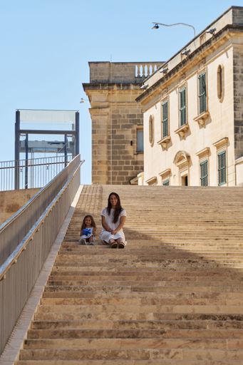 A vertical shot of a mother and her daughter sitting on a staircase with buildings and a blue sky in the background.