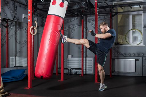 Young strong bearded mixed martial arts fighter trains in gym. Delivers powerful straight kick to red punching bag.