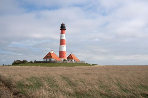 A grass field with a lighthouse and houses in Westerhever, Germany and a cloudy sky in the background.