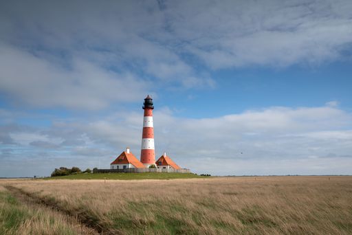 A grass field with a lighthouse and houses in Westerhever, Germany and a cloudy sky in the background.