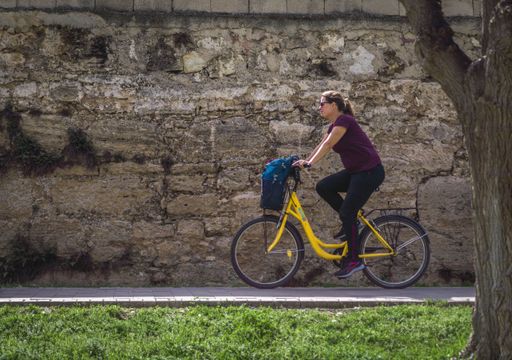 Picture of Urban Cyclist in Sport Tracksuit and Sunglasses Pedaling with her Yellow Bicycle through a Park with Green Grass and the Wall in the Backg