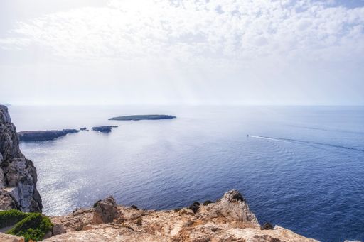 views of the cliffs and the mediterranean sea from the lighthouse of Cavalleria in Menorca, a boat leaves a wake on the waters on a summer day