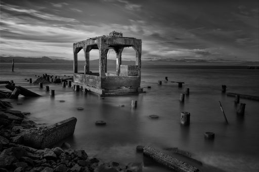 A greyscale shot of building ruins surrounded by wooden logs in the sea under the beautiful cloudy sky