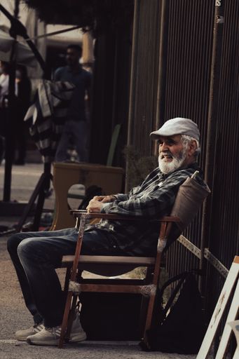 The vertical shot of a mature man sitting in front of a fence selling his artworks in Athens