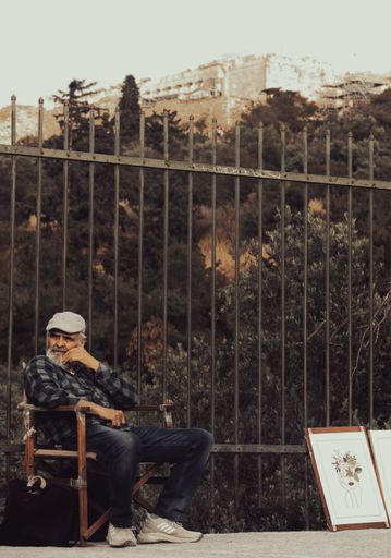 The vertical shot of a mature artist sitting in front of a fence selling his artworks in Athens
