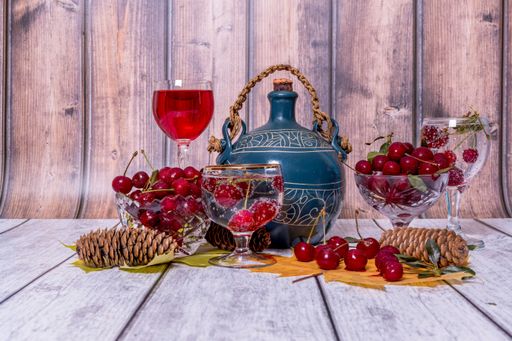 On wooden table are vases with ripe cherries, jug and glass of wine, glass of mineral water, yellow wedge leaves and fir cones.