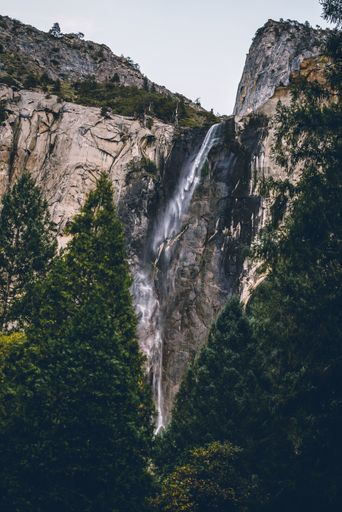 A vertical shot of a small waterfall inside high rocks and tall green trees in the front
