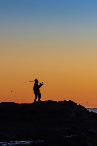 A vertical shot of a silhouette of a fisherman with his fishing rod at sunrise on the rocks of the sea