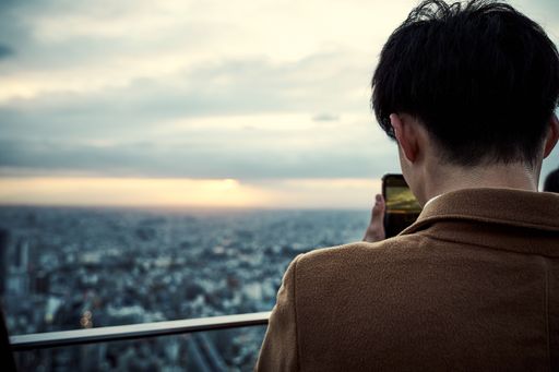 A closeup shot from the back of a man in balcony of a high building taking a cityscape picture with his phone