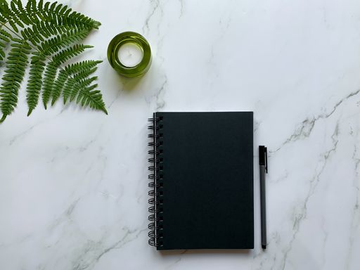 The flat lay of a black notepad, pen, and candle on a marble surface