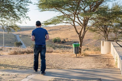 A male standing near the trees and looking at the road and the hills on a sunny day