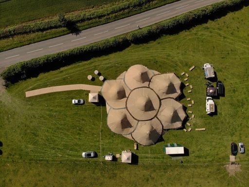 A bird's eye view of a flower shaped large tent in a green wide field along the highway