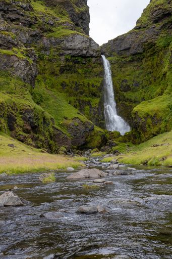 A vertical shot of a beautiful waterfall in Iceland surrounded by green hills and mossy rocks on a sunny day