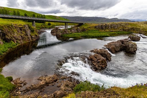A bridge built over a river in Iceland surrounded by gorgeous green hills and fields under a cloudy sky
