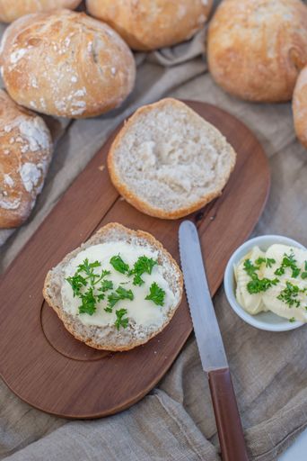 A vertical shot of a cut bread roll with butter and herbs next to other bread rolls in a basket