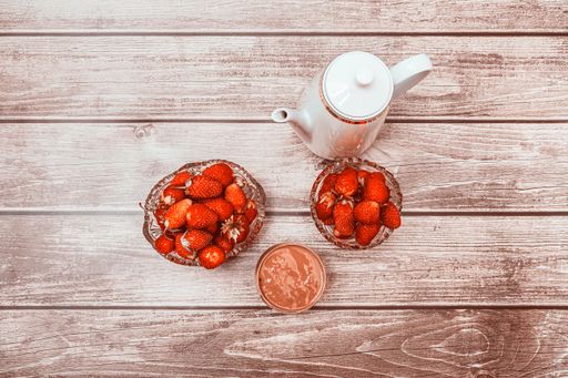 Two crystal vases filled with ripe strawberries, white coffee pot and cocktail glass stand on wooden surface. View from above. Tasty food.