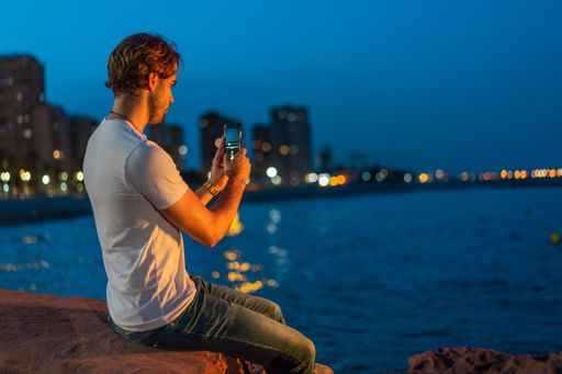 Caucasian man with casual clothes sitting on a stone of the boardwalk taking a photo with his cell phone at night.