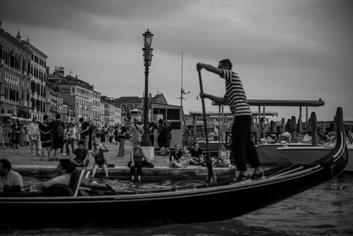 A greyscale shot of a man riding a gondola in Venice and many people walking happily in the streets