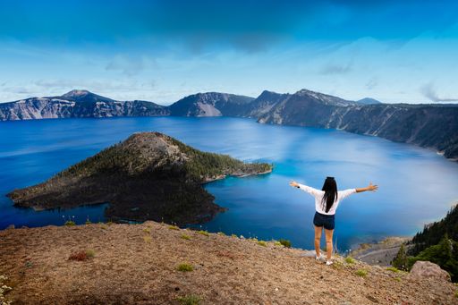 A high angle shot of a black-haired female standing on a hill and admiring the view of Crater lake in the United States