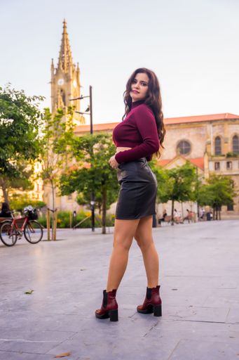 A vertical shot of a young stylish brunette female in leather skirt and a turtleneck posing in a city