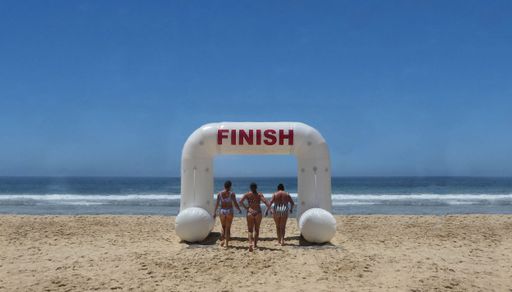 rear view of three women walking through inflatable finish archway on sand at the beach against clear blue sky