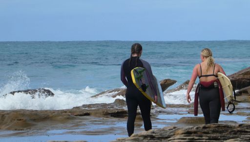 rear view of two young women wearing wetsuits carrying surfboards walking towards water going for a surf