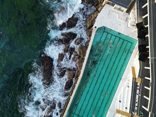 Aerial view of a swimming pool on a rocky coastline with resting people on a sunny day