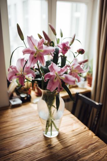 A vertical shot of lilies in a vase on a wooden table under the sunlight through the windows