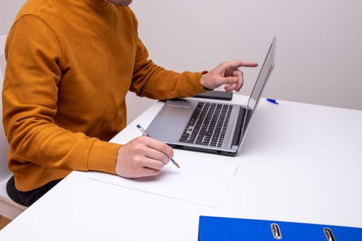A shot of a man sitting in front of a laptop while holding a pen to write something on a piece of paper