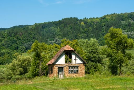 An old abandoned house in a field surrounded by hills covered in greenery under the sunlight