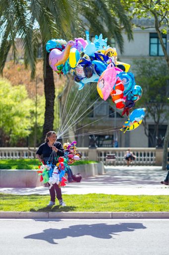 Barcelona / Spain - April 12 2015: Female Street Vendor Sells Animal Shaped Baloons in the Grass in a Public Park whit Palm Tree in the Background