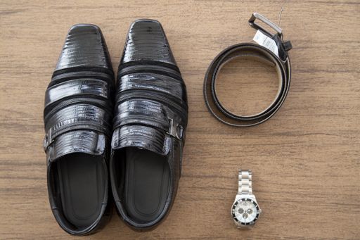 An overhead shot of a pairs of male black shoes and a belt with a watch on a brown surface