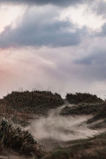 A vertical shot of flying sand over sand dunes with the cloudy sky in the background