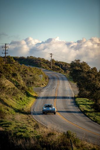 A vertical of a vintage American car driving along a mountain ridge with clouds and afternoon lighting