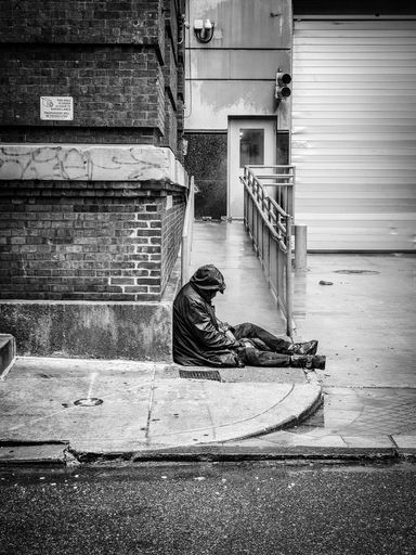 A vertical black and white shot of a homeless man sitting on the sidewalk near an apartment building